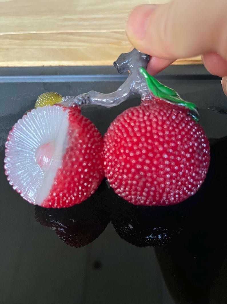 以下が含まれることがあります： 赤いライチの実2つのクローズアップ。1つはカットされ、白い果肉と種が見える。実はざらざらした表面で、緑の葉が付いた灰色の茎に付いている。ライチは手で持たれている。