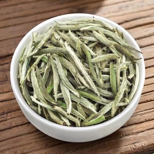 May include: A white ceramic bowl filled with dried silver needle white tea leaves. The tea leaves are long, slender, and covered in fine, pale silvery-green hairs, showcasing their natural, unprocessed state in a dense, textured pile.
