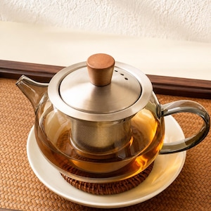 May include: A clear glass teapot featuring a brushed stainless steel lid with a small wooden knob, a matching stainless steel mesh infuser, and a curved glass handle. The teapot contains amber-colored liquid and sits on a round woven coaster.