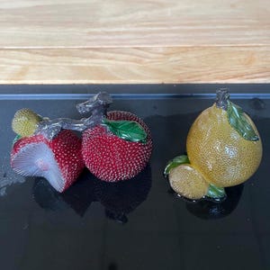 以下が含まれることがあります： 2つの装飾的なフルーツ彫刻：銀色の枝が付いた赤色のテクスチャ加工されたイチゴのペアと、緑の葉が付いた黄色のテクスチャ加工されたレモン。 フルーツは黒い表面にあります。