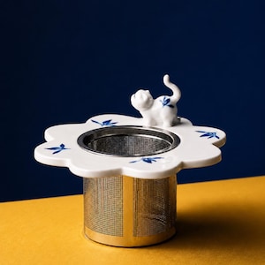 May include: A white ceramic tea infuser featuring a flower-shaped rim with blue leaf patterns and a small white cat figurine with blue markings on its back. The infuser has a cylindrical stainless steel mesh basket for steeping loose leaf tea.