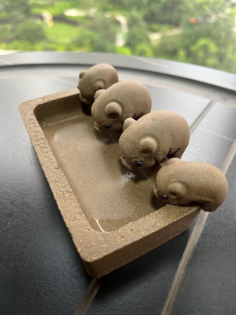 May include: A brown ceramic tray with four small, brown pig figurines. The tray has a rectangular shape with slightly raised edges. The pigs are positioned inside the tray, appearing to be drinking water. The background is blurred, suggesting an outdoor setting.