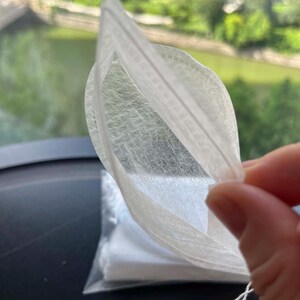 K&ouml;nnte beinhalten: Ein wei&szlig;er, halbtransparenter, rechteckiger Teebeutel aus d&uuml;nnem, faserigem Material. Der Beutel verf&uuml;gt &uuml;ber eine hei&szlig;versiegelte Kante und eine kleine wei&szlig;e Schnur an der unteren Ecke. Ein Stapel identischer Teebeutel ist darunter sichtbar.