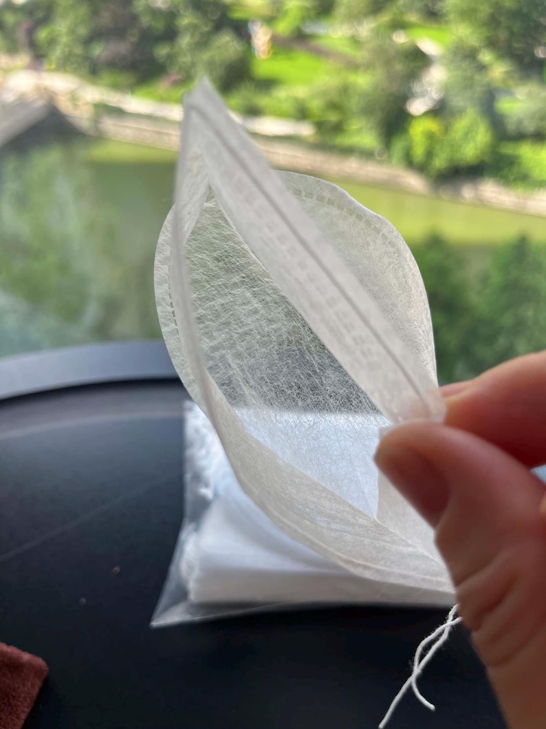 K&ouml;nnte beinhalten: Ein wei&szlig;er, halbtransparenter, rechteckiger Teebeutel aus d&uuml;nnem, faserigem Material. Der Beutel verf&uuml;gt &uuml;ber eine hei&szlig;versiegelte Kante und eine kleine wei&szlig;e Schnur an der unteren Ecke. Ein Stapel identischer Teebeutel ist darunter sichtbar.