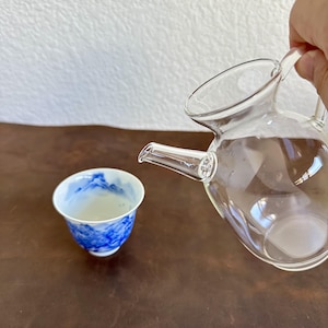 May include: A clear glass teapot with a curved handle and spout positioned next to a small white ceramic teacup featuring a blue painted landscape design on the interior and exterior surfaces.