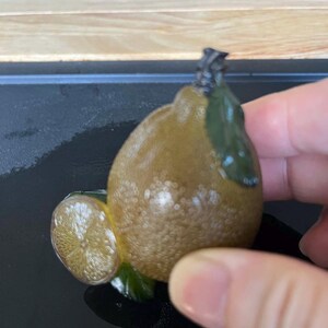以下が含まれることがあります： テクスチャ加工された茶色のボディと緑の葉が特徴の、スライス付きの装飾的なレモンの置物。レモンスライスは詳細な内部を明らかにします。置物は手で持たれています。