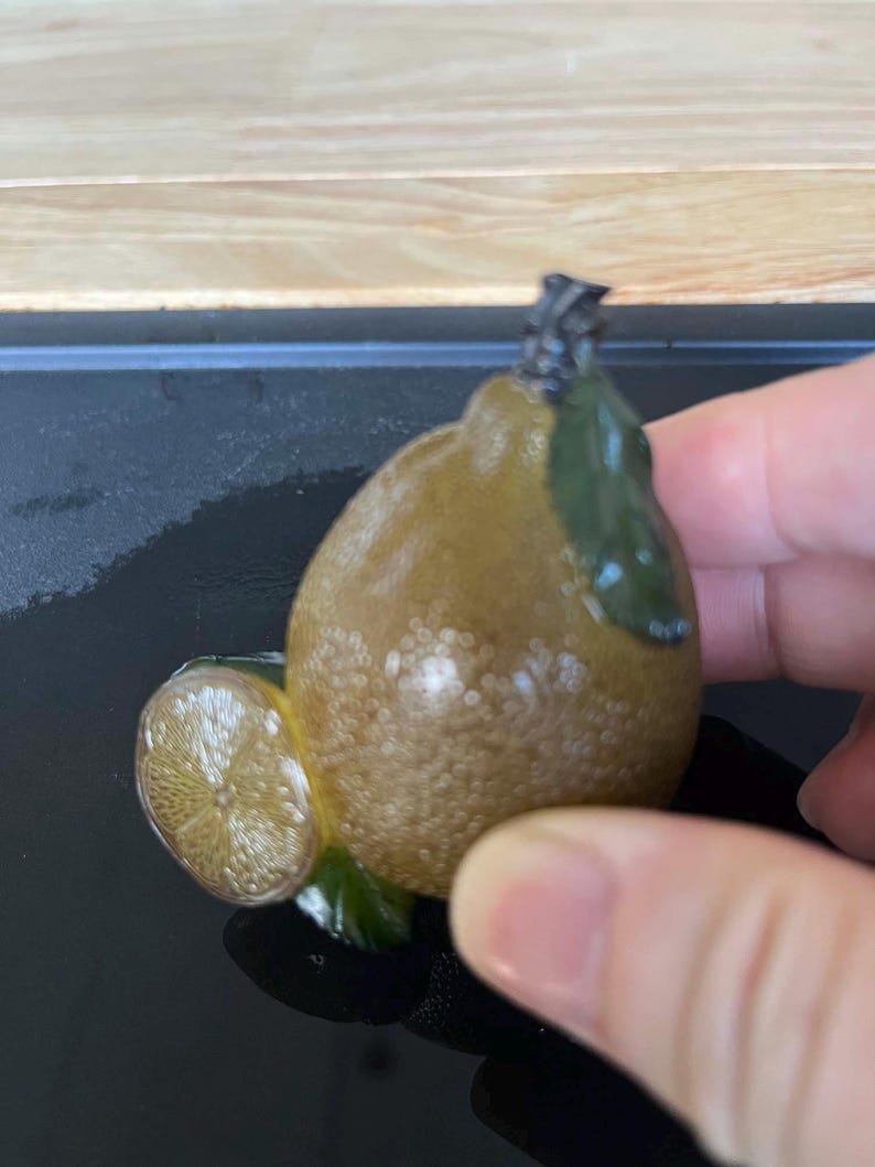 以下が含まれることがあります： テクスチャ加工された茶色のボディと緑の葉が特徴の、スライス付きの装飾的なレモンの置物。レモンスライスは詳細な内部を明らかにします。置物は手で持たれています。