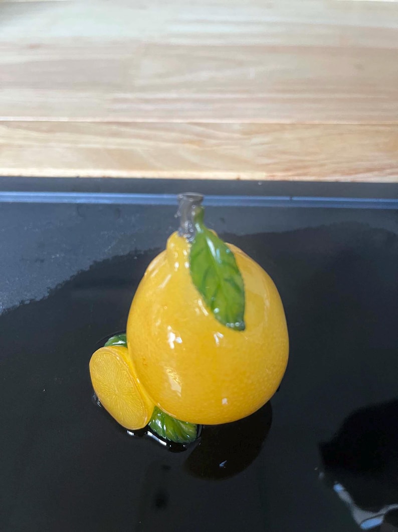 以下が含まれることがあります： 光沢のある黄色い仕上げ、緑の葉、スライスが特徴のセラミックレモンフィギュア。 レモンは黒い表面にあり、木製の背景があります。 このフィギュアは装飾品です。