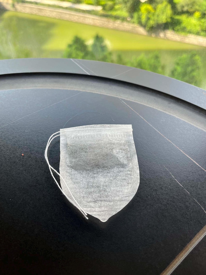 K&ouml;nnte beinhalten: Ein wei&szlig;er, halbtransparenter, rechteckiger Teebeutel mit abgerundeten unteren Ecken und einer d&uuml;nnen wei&szlig;en Schnur an der Oberkante. Der Teebeutel besteht aus einem feinen, por&ouml;sen Papiermaterial und enth&auml;lt lose Teebl&auml;tter im Inneren.