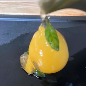 以下が含まれることがあります： 緑の葉が付いた、黄色のレモン型の装飾品。光沢があり、セラミックまたはガラス製のように見えます。目に見えないところからレモンに水が注がれています。