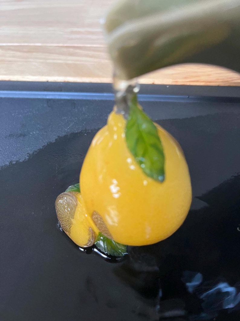 以下が含まれることがあります： 緑の葉が付いた、黄色のレモン型の装飾品。光沢があり、セラミックまたはガラス製のように見えます。目に見えないところからレモンに水が注がれています。