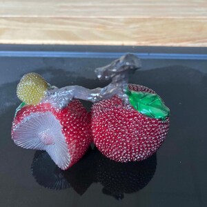 以下が含まれることがあります： 緑の葉と銀色の茎が付いた、2つの赤くテクスチャ加工されたベリーが特徴の装飾的なフルーツ彫刻。 1つのベリーはカットされており、詳細な内部が明らかになっています。彫刻は反射面を持っています。