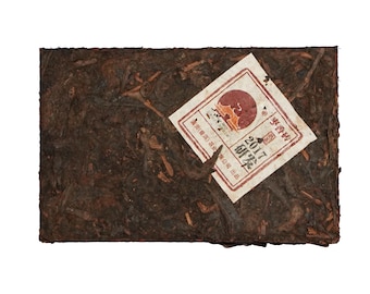 May include: A rectangular block of compressed tea, dark brown in color, with visible tea leaves and stems. A white and red label is affixed to the tea, with Chinese characters and the year "2017" printed on it. The tea appears to be a traditional, aged variety.
