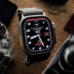 Könnte beinhalten: Eine schwarze Smartwatch mit schwarzem Armband, mit einem schwarzen Zifferblatt mit weißen Ziffern und Zeigern. Das Zifferblatt zeigt den Text "BLACKHAWK FLIEGER" und das Datum "04". Die Uhr hat ein graues Armband und einen gelben Akzent an der Seite.
