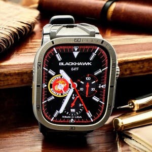 Op de afbeelding: Een vierkante smartwatch met een zwart display met de tekst "BLACKHAWK" en "64°F". De wijzerplaat heeft rode accenten, een Marine Corps-embleem en "Made in USA". Het horloge heeft een metallic grijze kast en een zwarte band.
