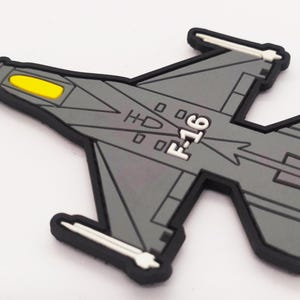 May include: A grey and black F-16 fighter jet patch with a yellow detail on the nose. The patch is made of rubber and has a textured surface.
