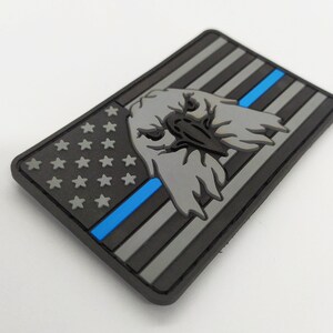 Peut inclure: Un patch noir et gris avec une bande bleue et une tête d'aigle grise. La tête d'aigle est tournée vers l'avant et a un bec. Le patch a un design qui ressemble au drapeau américain.