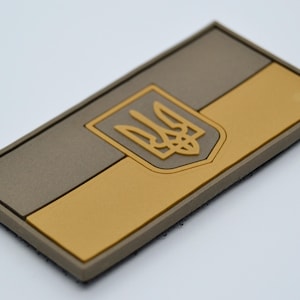 Könnte beinhalten: Ein brauner und gelber gestickter Aufnäher mit der ukrainischen Flagge und dem Staatswappen des Landes, einem Dreizack, in der Mitte.