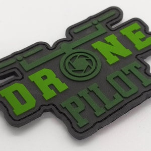 May include: A black and green patch with the words "DRONE PILOT" in bold green letters. The patch features a drone graphic and a dark green border. The design is a combination of green and black.