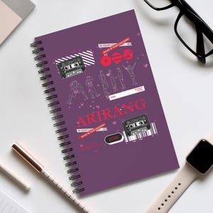 May include: A purple spiral-bound notebook featuring a graphic design with two black cassette tapes, red text reading ARIRANG, the year 2026, and the phrase WHAT IS YOUR LOVE SONG? The cover also includes line art of figures and small pink hearts.