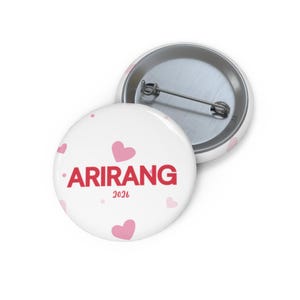 May include: White button pin with the word "ARIRANG" in red, accompanied by the year "2026" in smaller text. Pink hearts are scattered across the pin's surface. A second pin is partially visible.