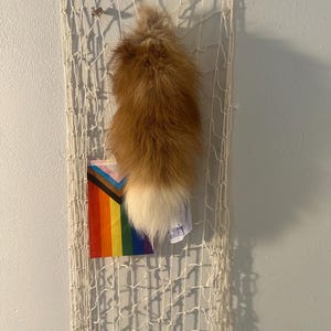 Könnte beinhalten: Eine dekorative Wandaufhängung aus geflochtenem weißem Seil mit einem flauschigen braun-weißen Fuchsschwanz. Eine Regenbogen-Pride-Flagge und Zeichnungen sind an der Aufhängung befestigt. Die Wand ist hellgrau gestrichen.