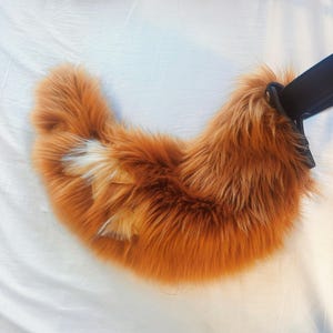 May include: A curved, plush faux fur tail in a vibrant orange hue with a distinct white tip. The tail is attached to a black leather-like strap with a metallic silver-toned snap button fastener at the base, creating a crescent-shaped accessory.