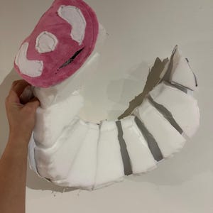 May include: A curved, segmented prop constructed from white foam pieces. The top section features a rounded, plush pink head covering with white fabric accents and a dark horizontal slit, creating a distinct, multi-part creature-like structure.