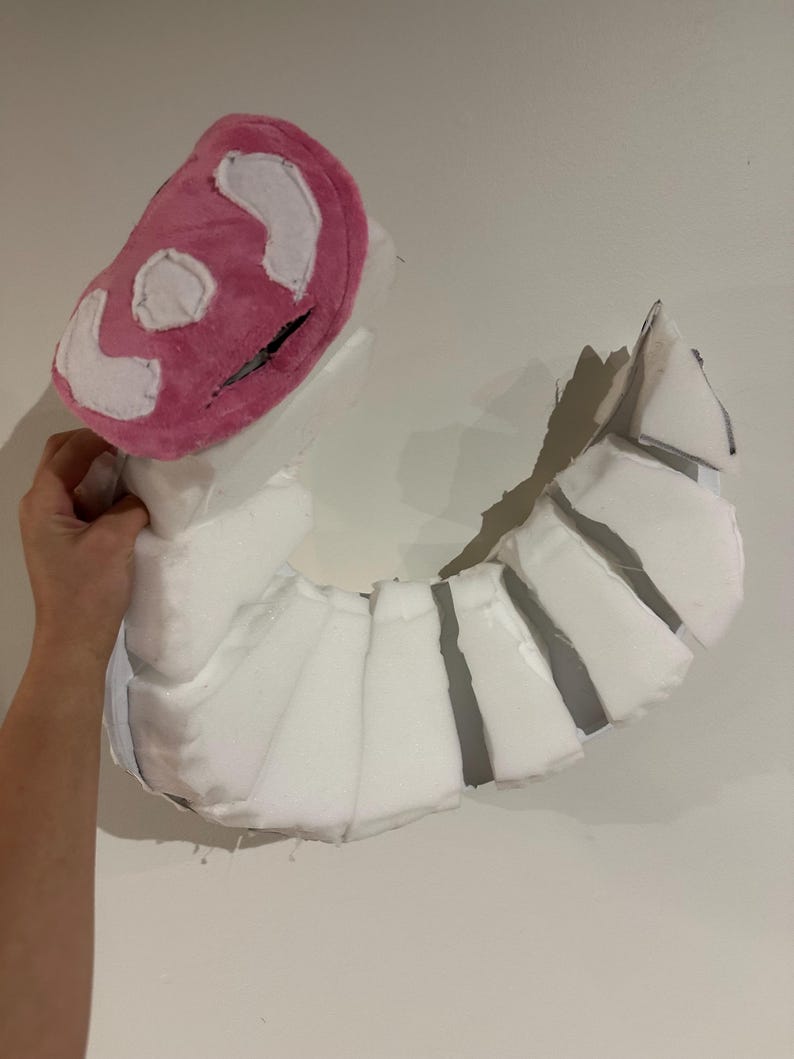 May include: A curved, segmented prop constructed from white foam pieces. The top section features a rounded, plush pink head covering with white fabric accents and a dark horizontal slit, creating a distinct, multi-part creature-like structure.