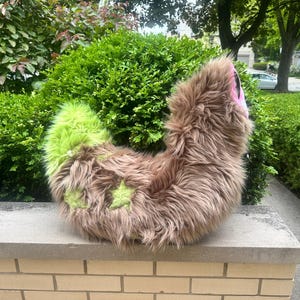 May include: A plush tail accessory made of long, shaggy brown faux fur with a bright lime green tip and two lime green star-shaped patches on the side. The tail features a curved, flexible design with a pink fabric accent visible at the top end.