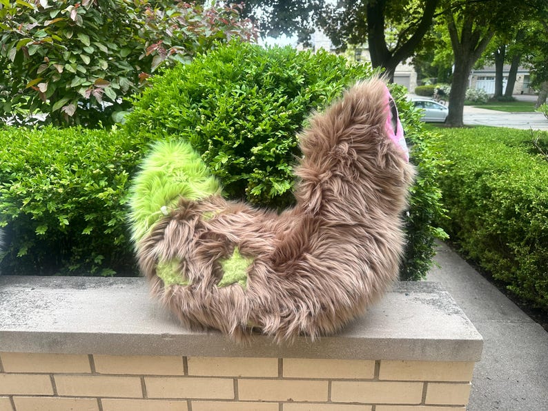 May include: A plush tail accessory made of long, shaggy brown faux fur with a bright lime green tip and two lime green star-shaped patches on the side. The tail features a curved, flexible design with a pink fabric accent visible at the top end.