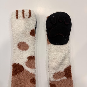 May include: A pair of cozy, fluffy socks with a cat paw design. The socks are white, brown, and tan, with a black paw pad on the sole. The socks are designed to resemble a cat's paw.
