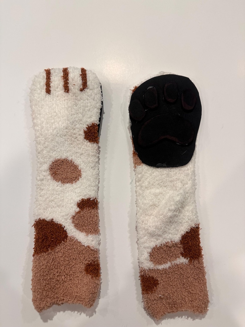 May include: A pair of cozy, fluffy socks with a cat paw design. The socks are white, brown, and tan, with a black paw pad on the sole. The socks are designed to resemble a cat's paw.