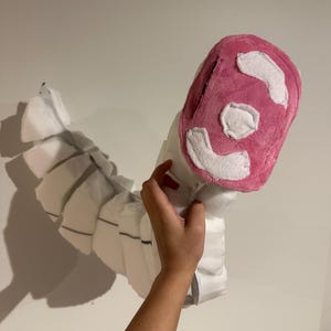 May include: A plush, segmented white tail accessory featuring a rounded, bright pink tip with white fabric patches. The long, curved structure is composed of multiple connected sections, creating a flexible, articulated appearance for costume use.