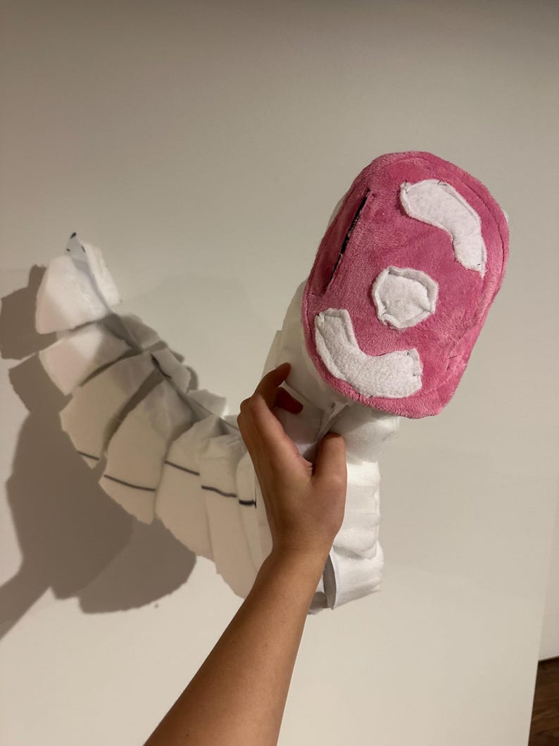 May include: A plush, segmented white tail accessory featuring a rounded, bright pink tip with white fabric patches. The long, curved structure is composed of multiple connected sections, creating a flexible, articulated appearance for costume use.