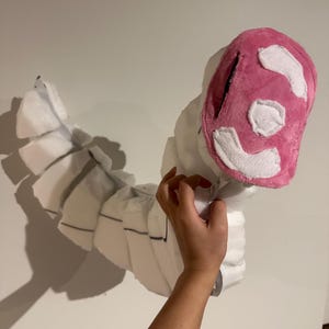 May include: A segmented white plush creature with a curved, flexible body and a pink, oval-shaped head. The head features white fabric patches arranged in a pattern. The body consists of multiple white foam-like sections joined together.