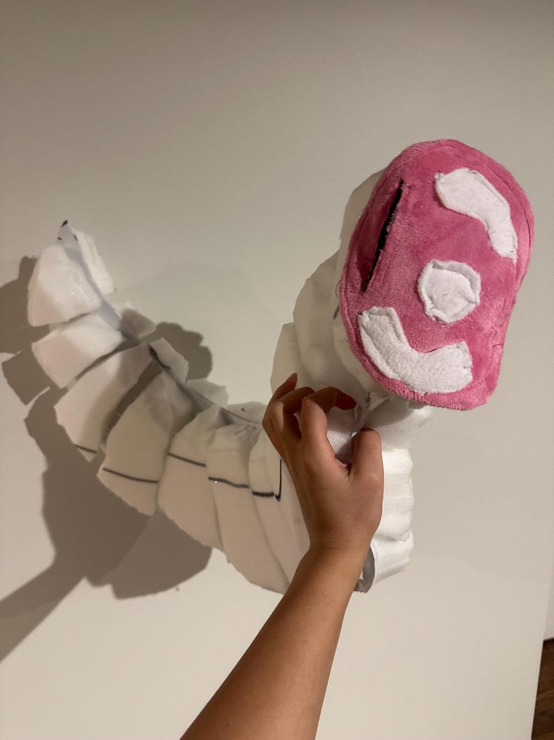 May include: A segmented white plush creature with a curved, flexible body and a pink, oval-shaped head. The head features white fabric patches arranged in a pattern. The body consists of multiple white foam-like sections joined together.