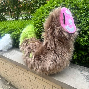 May include: A plush tail accessory made of long brown faux fur with a bright lime green section near the base. The tip of the tail features a rounded pink fabric patch with a white stitched design, creating a textured and soft appearance.
