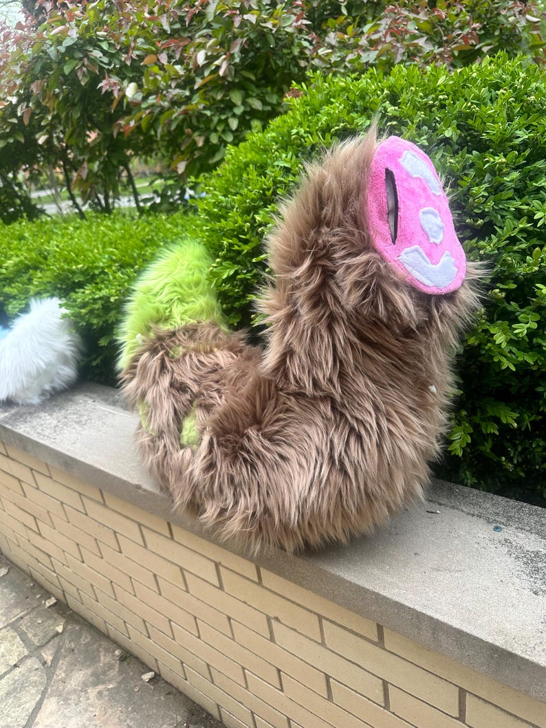 May include: A plush tail accessory made of long brown faux fur with a bright lime green section near the base. The tip of the tail features a rounded pink fabric patch with a white stitched design, creating a textured and soft appearance.