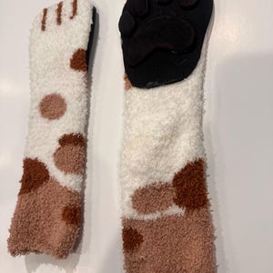 May include: Two fuzzy cat paw socks. One sock features a white and brown spotted pattern, while the other has a black paw pad design. The socks are made of a soft, plush material.