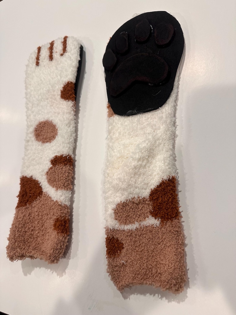 May include: Two fuzzy cat paw socks. One sock features a white and brown spotted pattern, while the other has a black paw pad design. The socks are made of a soft, plush material.