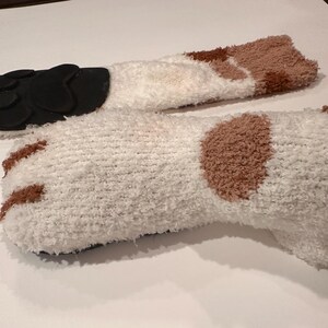 May include: A pair of cozy, white fuzzy socks with brown patches and black paw pads on the soles. The socks are designed to resemble a cat's paws, offering a playful and comfortable accessory.