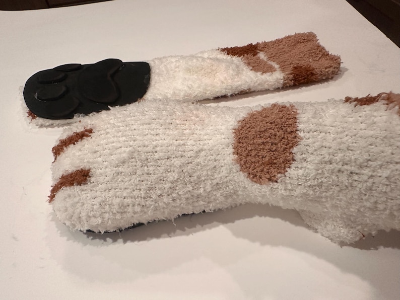 May include: A pair of cozy, white fuzzy socks with brown patches and black paw pads on the soles. The socks are designed to resemble a cat's paws, offering a playful and comfortable accessory.