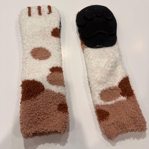 May include: A pair of cozy, fluffy socks with a cat paw design. The socks are white with brown spots and a brown toe area. The soles have a black paw print design.