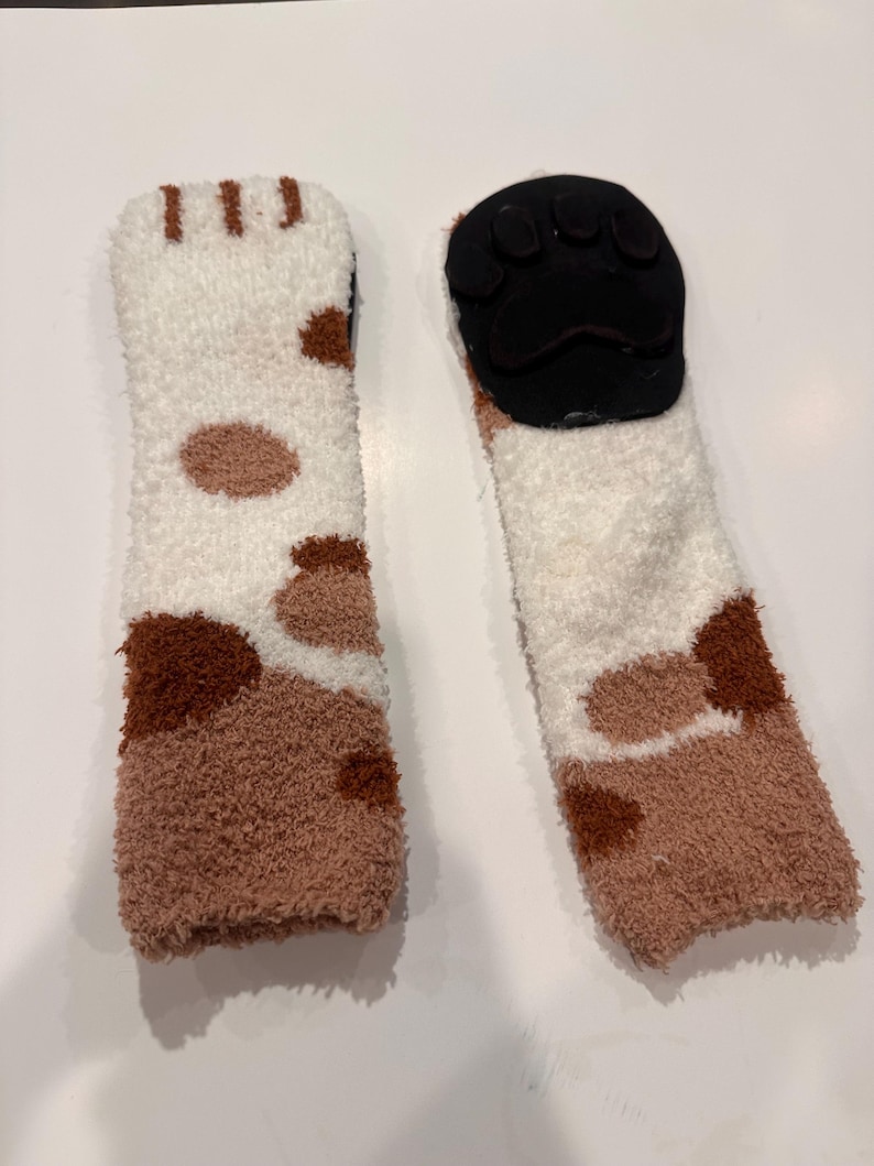 May include: A pair of cozy, fluffy socks with a cat paw design. The socks are white with brown spots and a brown toe area. The soles have a black paw print design.