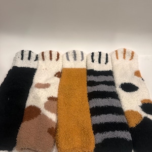 May include: Five pairs of fuzzy socks in various colors and patterns. The socks include black, white with brown spots, orange, black and gray stripes, and white with black and brown spots. Each pair has a striped cuff.