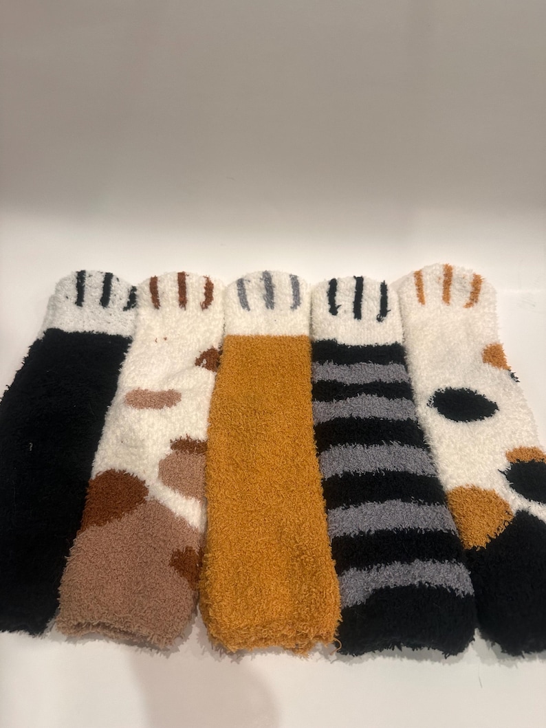 May include: Five pairs of fuzzy socks in various colors and patterns. The socks include black, white with brown spots, orange, black and gray stripes, and white with black and brown spots. Each pair has a striped cuff.