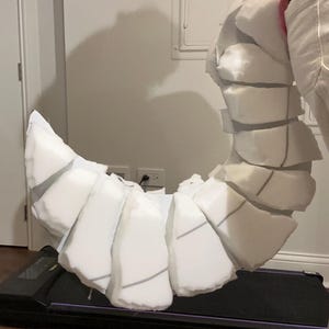 以下が含まれることがあります： 白のフォームで作られた、大きく湾曲した、セグメント化された小道具。小道具は黒い表面で支えられています。セグメントは灰色の線で縁取られています。小道具は尾または角のように見えます。