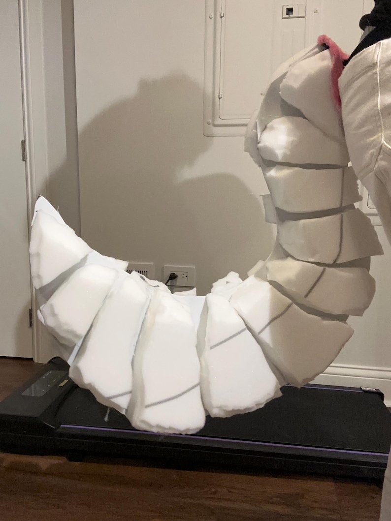 May include: A curved, segmented tail prop constructed from white foam blocks. The structure features multiple individual foam pieces arranged in a series to create a flexible, articulated appearance, with a small pink accent visible at the top attachment point.