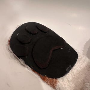 May include: A black dog paw-shaped item with three oval pads and a larger pad, set against a white and brown fluffy background. The paw is made of a dark, possibly felt-like material.